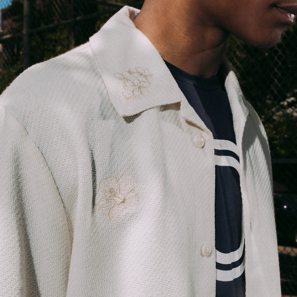 Close-up of a person wearing a beige collared shirt with floral embroidery over a black 'Overtime' tee