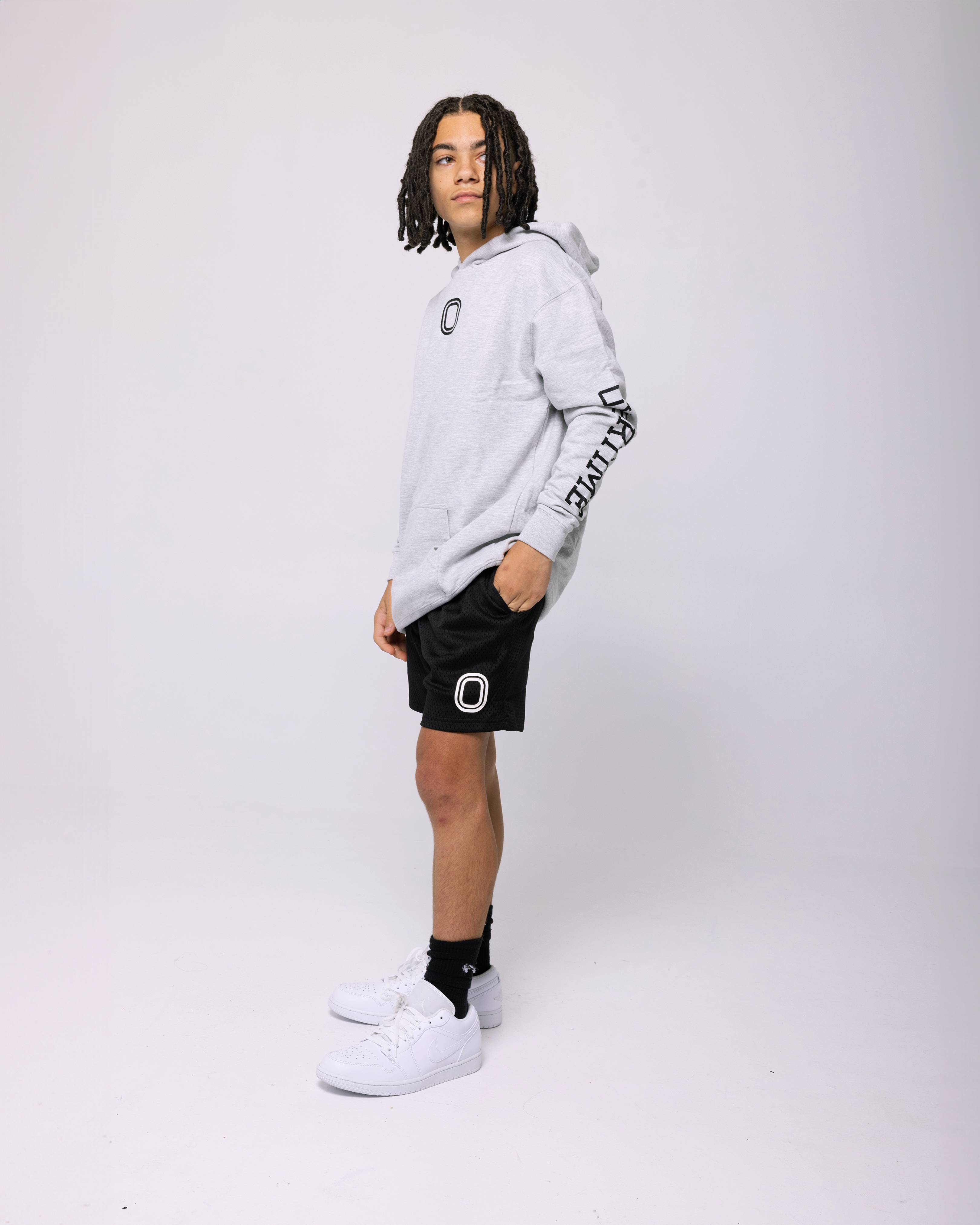 A kid wearing a grey hoodie with a 'O' logo print on the chest and 'OVERTIME' print on the sleeve.
