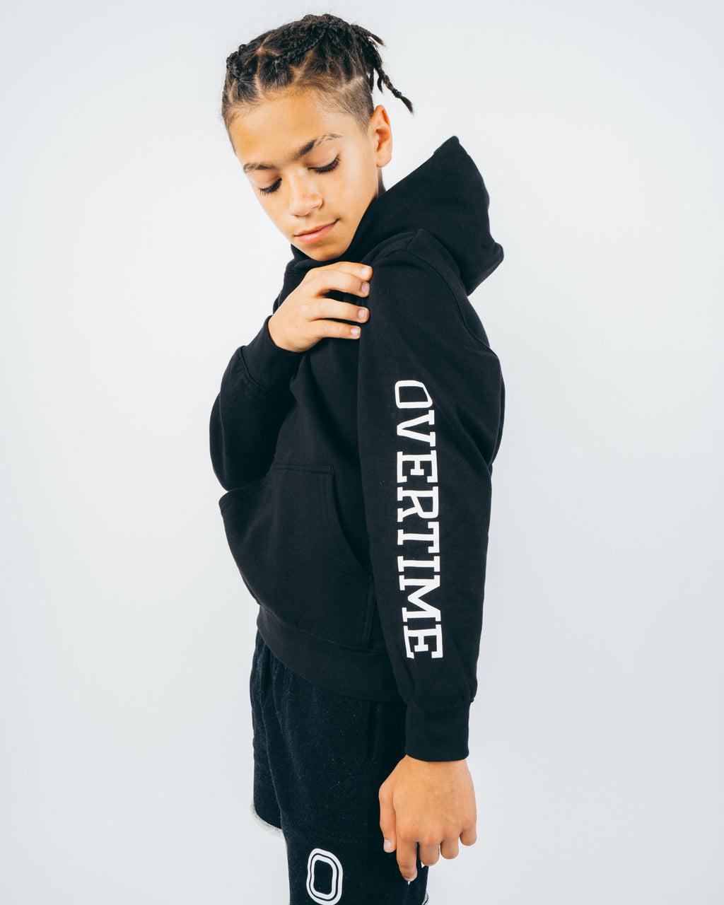 A kid wearing a black hoodie with a 'O' logo print on the chest and 'OVERTIME' print on the sleeve. He's showing off the sleeve. 