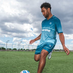 Man in blue sports outfit kicking a soccer ball on a field with a cloudy sky.