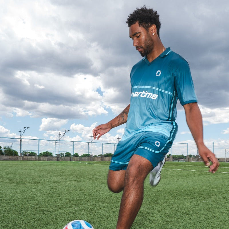 Man in blue sports outfit kicking a soccer ball on a field with a cloudy sky.