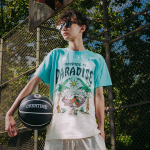 Person on a basketball court wearing a graphic tee that says 'Hooping is Paradise' and holding an Overtime basketball
