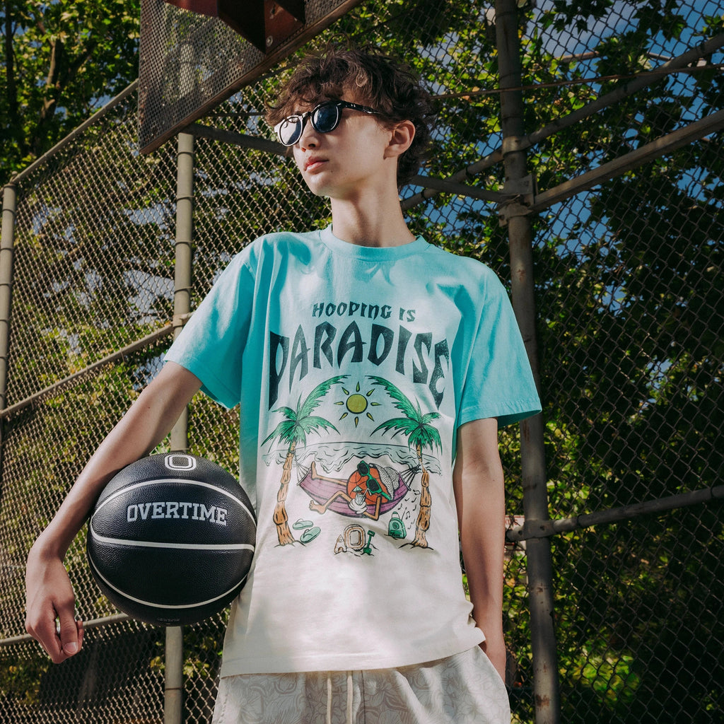 Person on a basketball court wearing a graphic tee that says 'Hooping is Paradise' and holding an Overtime basketball